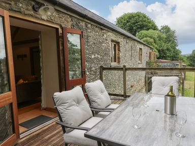 An outdoor seating area with cushioned chairs and a table with wine glasses and a bottle outside a stone building at Nant Glas Pumpsaint near Lampeter
