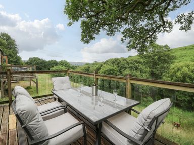 An outdoor wooden deck with a table and four chairs surrounded by trees and hills at Nant Glas in Pumpsaint near Lampeter