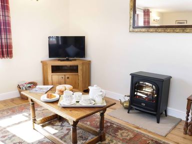 A living room with a wooden coffee table set with tea cups and cake near a television and electric stove at The Cider House in Leominster