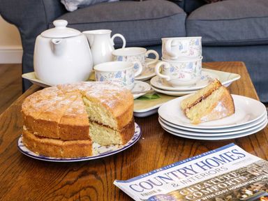 A wooden table with a sliced sponge cake tea set and a magazine in a living room at The Cider House in Leominster
