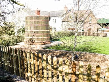 A wooden barrel structure in a garden with a wooden fence and houses in the background at The Cider House in Leominster