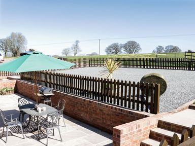 An outdoor patio with a table, chairs, green umbrella, and a barbecue grill next to a gravel area with a wooden fence and trees in the background at The Cider House in Leominster