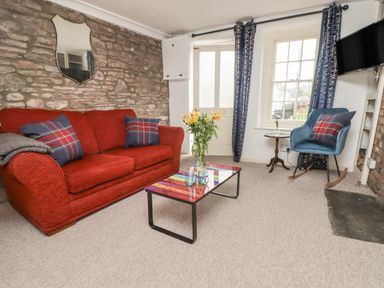 A living room with a red sofa and blue rocking chair next to a window and a multicolored coffee table with flowers at Kooky Cottage in Brecon