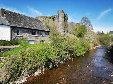 A stone house next to a river with trees and an old castle tower in the background at Kooky Cottage in Brecon