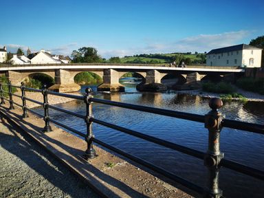 A stone bridge over a river with a metal railing in the foreground and houses and hills in the background at Kooky Cottage in Brecon