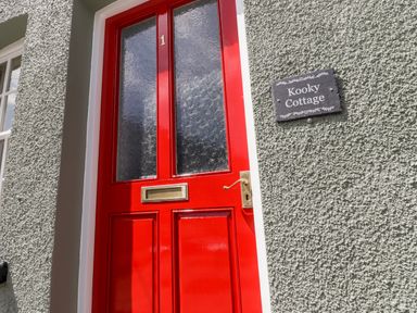 A red front door with the number 1 and letterbox on a textured grey wall with a Kooky Cottage sign in Brecon