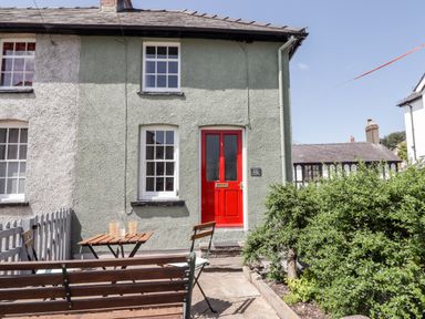 An exterior view of a house with a red door windows a wooden bench table and chairs and a green bush at Kooky Cottage in Brecon