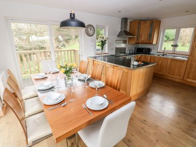 A kitchen and dining area with wooden cabinets a dining table set for six and large windows overlooking a garden at The Dingle in Wisemans Bridge near Stepaside