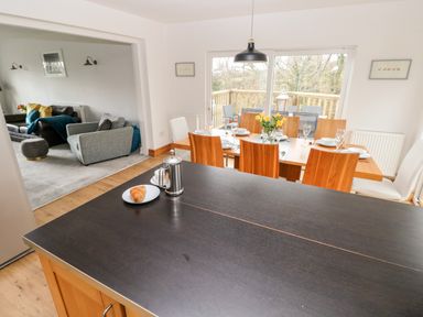 A dining area with a wooden table set for six and a vase of yellow flowers next to a kitchen island with a croissant and coffee pot and a living room with sofas in the background at The Dingle in Wisemans Bridge near Stepaside