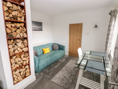 A room with a teal sofa with cushions a glass table with a white chair stacked firewood in shelves and a wooden door at The Dingle in Wisemans Bridge near Stepaside