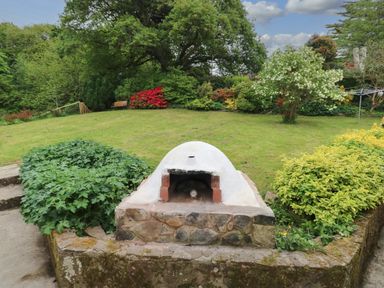 A garden with a stone oven in the foreground surrounded by bushes and trees at The Dingle in Wisemans Bridge near Stepaside