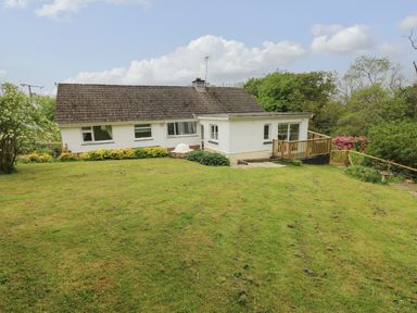A white house with multiple windows and a wooden deck surrounded by grass and trees at The Dingle in Wisemans Bridge near Stepaside