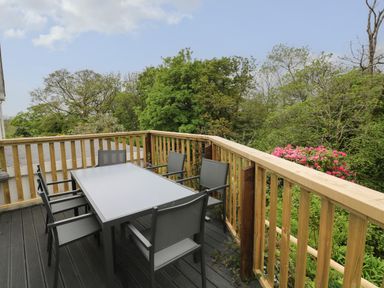 An outdoor deck with a table and six chairs surrounded by wooden railings and trees at The Dingle in Wisemans Bridge near Stepaside