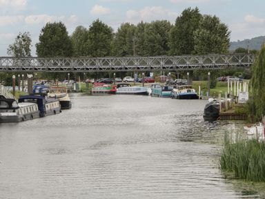 A river with boats docked along both sides and a metal footbridge crossing over the water at Runnymede in Tewkesbury