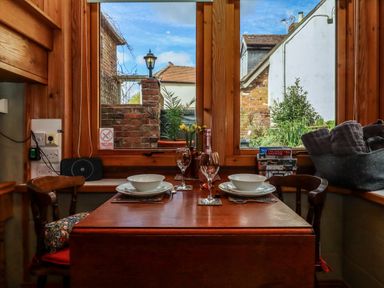 A dining table set for two with bowls wine glasses flowers and a bottle by a window with a view of a brick wall and garden at Runnymede in Tewkesbury