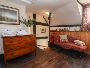 A corner room with a wooden chest of drawers with towels and a vase on top and a red patterned chaise lounge with cushions and a rolled blanket at Runnymede in Tewkesbury