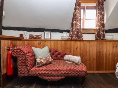 A room with a patterned red chaise lounge near a wooden paneled wall with curtains on a window at Runnymede in Tewkesbury