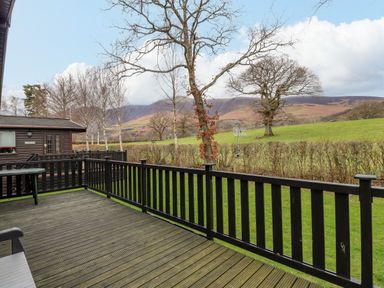 A wooden deck with black railings overlooking a grassy field with trees and hills at The Fold Number 28 Burnside Park near Keswick