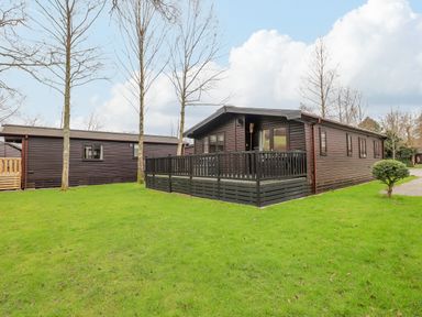 A wooden cabin with a fenced porch on a grassy lawn with trees at The Fold in Number 28 Burnside Park near Keswick