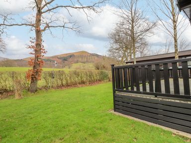 A grassy garden area with trees and a black wooden deck overlooking hills at The Fold Number 28 Burnside Park near Keswick