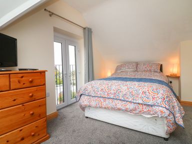 A bedroom with a double bed with patterned bedding a wooden dresser a wall-mounted television and French doors at Hill Coach House in Watchet