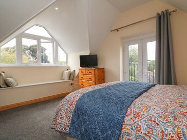 A bedroom with a bed a wooden chest of drawers a window seat with cushions and French doors with curtains at Hill Coach House Watchet
