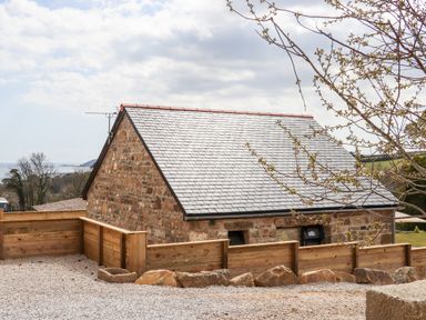 A stone house with a slate roof and wooden fence outside with a view of trees and sea at Apple Blossom Barn in Gulval near Penzance