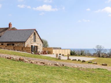 A stone house with a slate roof and outdoor seating area on a grassy slope with a view of the sea at Apple Blossom Barn in Gulval near Penzance