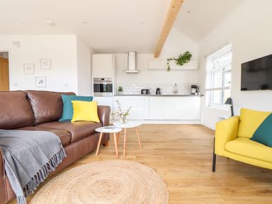 A living area with brown and yellow sofas two small white tables with plants and books a kitchen with white cabinets and appliances at Apple Blossom Barn in Gulval near Penzance