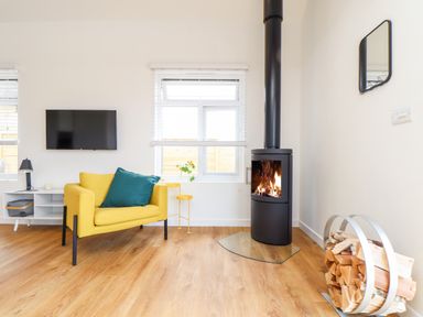 A living room with a yellow armchair a black wood stove and firewood at Apple Blossom Barn Gulval near Penzance