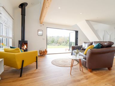 A living room with a yellow armchair a brown leather sofa a round rug a wood burning stove and an open glass door at Apple Blossom Barn in Gulval near Penzance