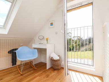 A small corner with a blue rocking chair next to a white desk and an open door overlooking a grassy garden at Apple Blossom Barn in Gulval near Penzance
