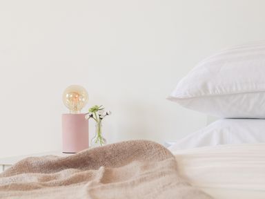 A bedside table with a pink lamp and small vase with flowers next to a bed with white pillows and a beige blanket at Apple Blossom Barn in Gulval near Penzance