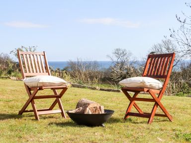 Two wooden chairs with cushions around a fire pit on grass with sea and trees in the background at Apple Blossom Barn in Gulval near Penzance