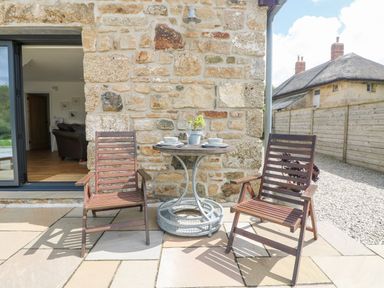 An outdoor seating area with two wooden chairs and a round table set against a stone wall at Apple Blossom Barn in Gulval near Penzance