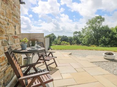 A patio with two wooden chairs and a table set with plates and glasses next to a stone wall overlooking a garden with grass and trees at Apple Blossom Barn in Gulval near Penzance