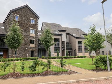 Modern residential buildings with stone and grey walls and multiple windows surrounded by green grass and small trees at Rivers Edge in Newby Bridge