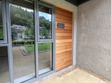 The entrance to a property with a wooden door numbered 24 and large glass windows reflecting trees and nearby buildings at Rivers Edge in Newby Bridge