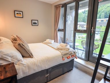 A bedroom with a made bed and towels on top next to a large window with an outdoor seating area at Rivers Edge in Newby Bridge