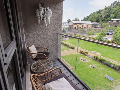 A balcony with two wicker chairs with cushions and a glass-top table overlooking a grassy area with buildings in the background at Rivers Edge in Newby Bridge