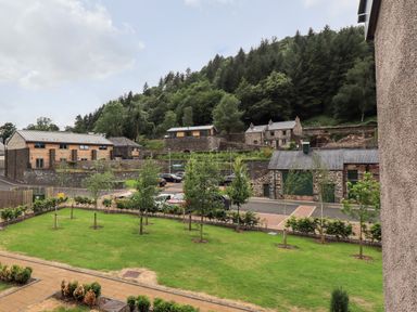 A grassy yard with small trees and bushes bordered by a sidewalk with stone and timber houses and a parking area in front of a wooded hill at Rivers Edge in Newby Bridge