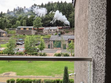A view of a grassy area with trees cars and stone buildings with a steam train passing by in the background at Rivers Edge in Newby Bridge