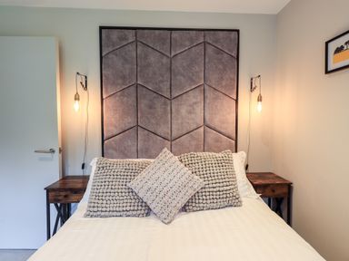A bedroom with a bed with three pillows and a large padded headboard with two bedside tables and hanging light bulbs at Rivers Edge in Newby Bridge