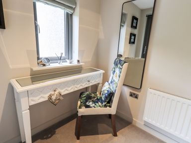 A white desk with decorative items by a window and a floral upholstered chair next to a tall wall mirror at Rivers Edge in Newby Bridge