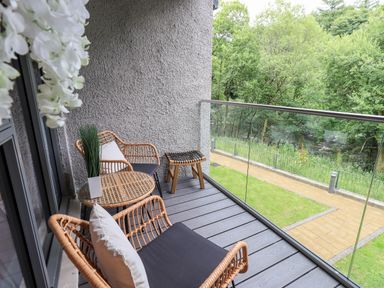 A balcony with wicker chairs a small table and a stool overlooking a path and trees at Rivers Edge in Newby Bridge