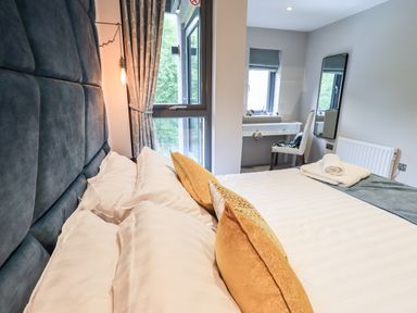 A bedroom with a bed featuring white pillows and gold cushions next to a window with curtains and a vanity desk with a chair at Rivers Edge in Newby Bridge