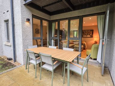An outdoor patio with a wooden table and six chairs next to a building with large windows and an open door showing a living room inside at Rivers Edge in Newby Bridge