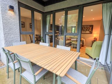 An outdoor patio area with a wooden table and six chairs beside large glass doors showing a living room with an armchair sofa and wall art at Rivers Edge in Newby Bridge