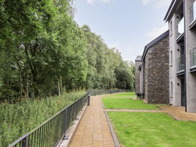 A paved walkway beside grass and a row of trees next to stone and glass buildings at Rivers Edge in Newby Bridge