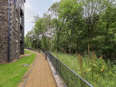 A paved walkway with a stone building on the left and trees with greenery on the right at Rivers Edge in Newby Bridge